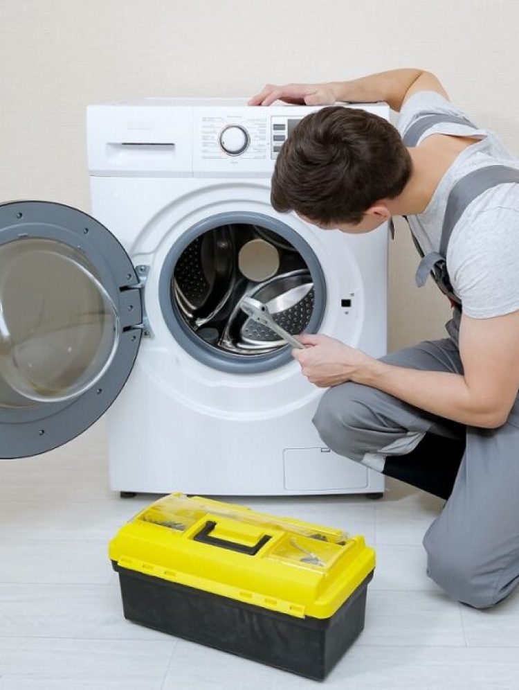 young serviceman in grey uniform repairs broken washing machine with wrench on wooden floor near beige wall